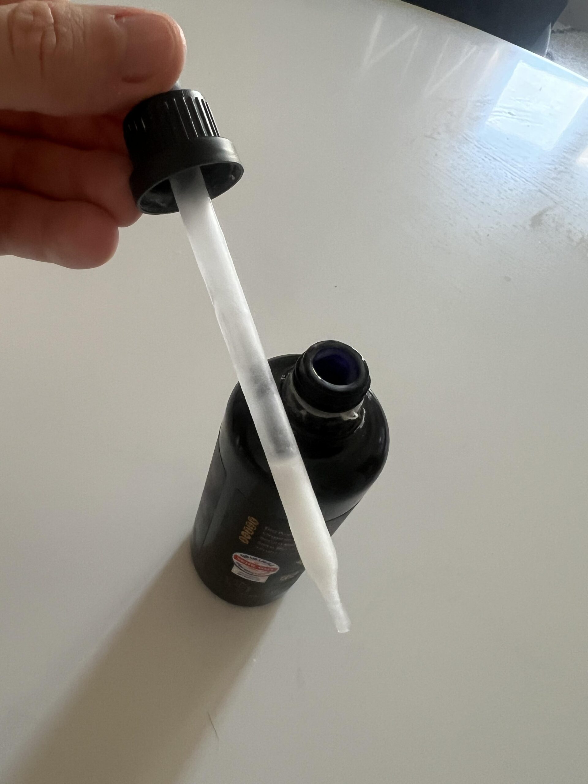 My original photo of the transparent pipette with pale Kapua Naturals azelaic acid serum inside, held over the open black bottle